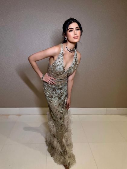 A full-length shot of a glamorous evening look. The makeup is soft and glowing, with a focus on flawless skin, complementing the sequined gown with a feather-trimmed hem.