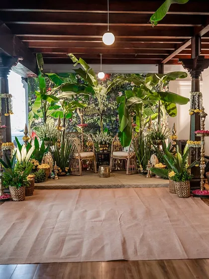 The main stage for a Haldi ceremony, set against a lush tropical backdrop. The seating is flanked by banana plants, brass lamps, and baskets of flowers, rooting the design in nature and tradition.