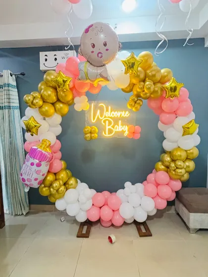 This is a modern and stylish setup for a baby shower. It features a circular balloon wreath in pink, white, and gold, with a 'Welcome Baby' neon sign that looks great in photos.