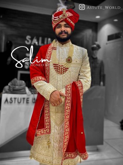 A classic groom's ensemble featuring an ivory sherwani with gold embroidery. The look is completed with a deep red safa and a matching red velvet stole, creating a timeless and regal combination.