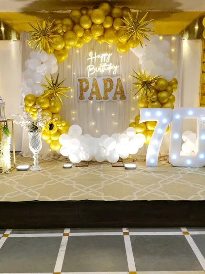We create elegant setups for milestone celebrations, like this 70th birthday party. The decor features a classic gold and white balloon arch, a sheer curtain backdrop with fairy lights, a custom neon sign, and a large "70" marquee number, creating a sophisticated and celebratory atmosphere.