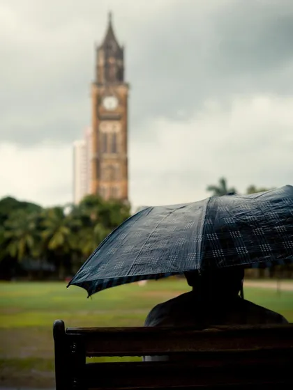 A person sits on a bench, silhouetted under an umbrella, with the Rajabai Clock Tower blurred in the background. The shallow depth of field creates a moody and mysterious portrait.