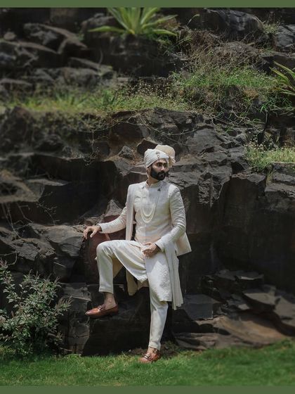 A powerful portrait of the groom in his ivory sherwani, posing against a natural rock formation. The composition is strong and regal, befitting a groom on his wedding day.