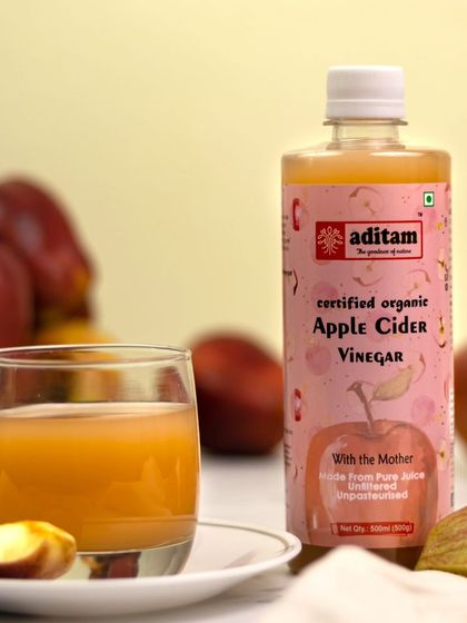 A bottle of certified organic apple cider vinegar is styled with fresh apples and a glass of the diluted drink. The warm, natural lighting and simple setup highlight its wholesome, healthy qualities.