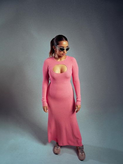 A full-length shot of a contemporary pink dress. The minimalist studio setting and the model's confident stance highlight the dress's unique cut-out design and modern silhouette.