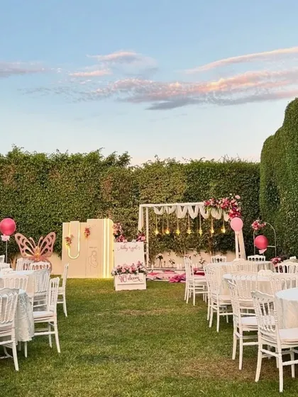 A beautiful outdoor sundowner setup for a 'Welcome Baby Girl' party, with elegant white tables, a low-seating area, and multiple themed photo backdrops.