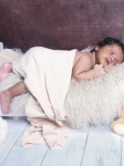 A classic sleeping pose on a miniature bed, with a teddy bear companion. The simple white wood floor and blanket keep the focus on the peaceful baby.