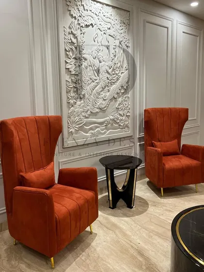 A pair of high-back chairs in a burnt orange velvet, set against a wall with intricate white molding. The chairs' tall, slender profile and rich color add a sense of drama and luxury.