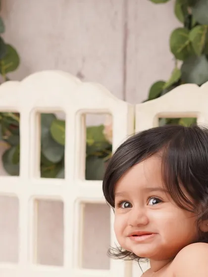 A sweet close-up portrait of a baby girl with an endearing expression, framed by a rustic white window prop and a leafy garland.