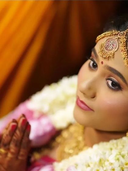 A beautiful candid photo from Navya's wedding ceremony. This shot captures her serene expression and the flawless finish of her traditional bridal makeup, complete with gold jewellery and a flower garland.