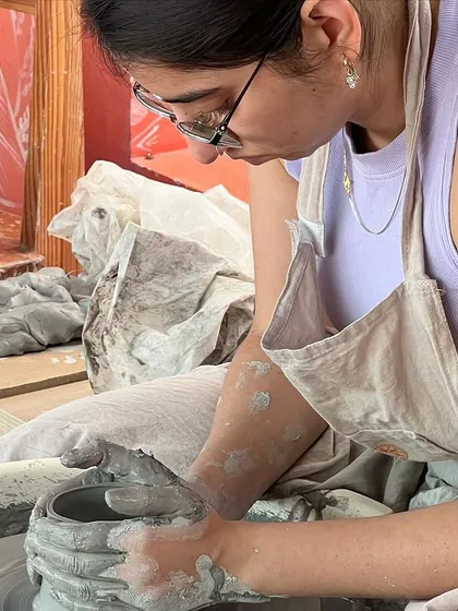 A close-up of a student's hands, covered in clay, working to shape a pot on the wheel. This is what it's all about.