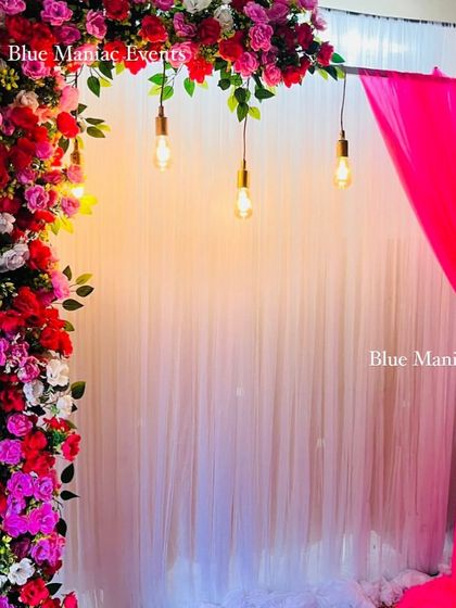An engagement or anniversary backdrop with a floral arch and hanging Edison bulbs. The mix of pink, red, and white flowers creates a romantic and vibrant feel.