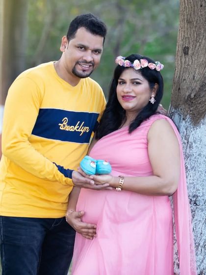 A smiling couple holds up a pair of tiny blue baby shoes, announcing they are expecting a boy. The mother's pink dress and floral crown complete this happy portrait.