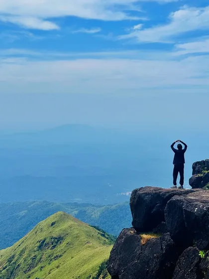 A trekker making a heart shape with his hands, overlooking the vast green valleys from a cliff on the Kumara Parvatha trail.