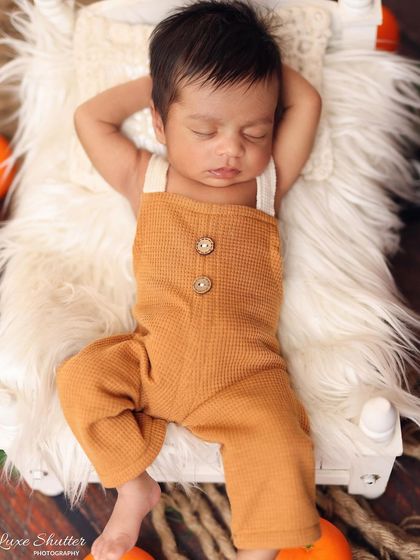 A relaxed and adorable pose of a newborn in cute overalls, sleeping on a fluffy rug. This shows a more natural, unswaddled style of newborn photography.