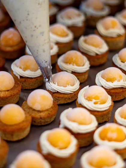 A mesmerizing close-up of mini cakes being prepared. A pastry bag pipes a perfect swirl of cream onto each one, showing the consistency and skill of our culinary team.