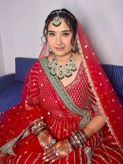 A full view of the bride seated, looking serene and beautiful. The makeup, featuring emerald green jewelry, adds a touch of regal elegance to her look.