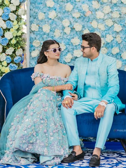 A stylish portrait of the couple seated on a royal blue sofa, set against a floral wall. Their coordinated outfits and cool sunglasses give this pre-wedding shot from Pushkar a modern, chic vibe.