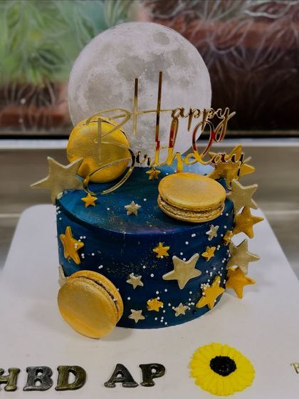 A cake that's out of this world! This galaxy-themed design features a moon topper, edible planets, stars, and macarons for a celestial birthday celebration.
