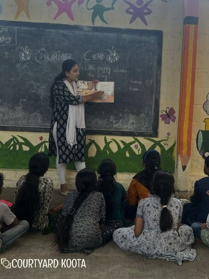 A storytelling session in progress at the Makkala Masti summer camp. A volunteer reads a picture book to an attentive group of children, fostering a love for reading and language.