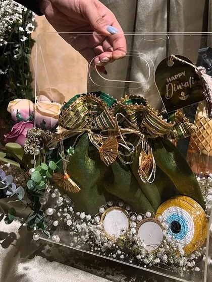 A stunning acrylic hamper for Diwali, featuring a potli filled with treats, an evil eye charm, and beautiful floral decorations.
