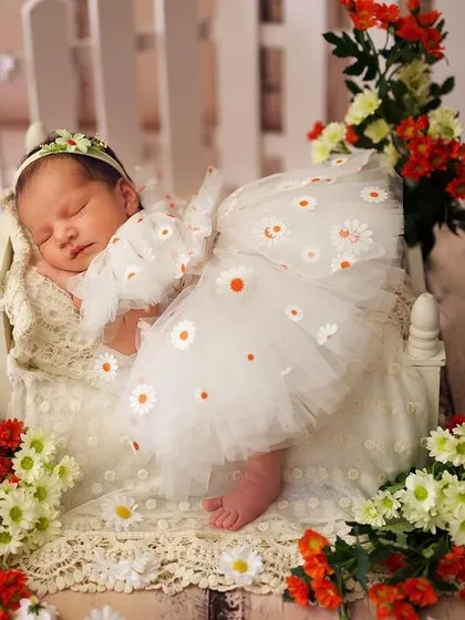 A dreamy garden scene with a baby in a daisy-print dress sleeping on a miniature bed, surrounded by fresh flowers.