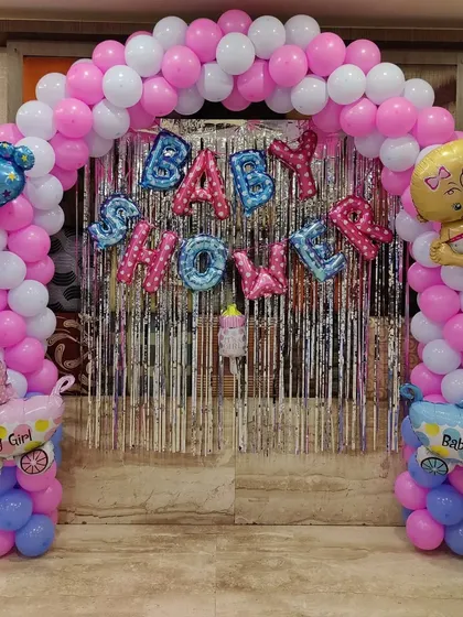 A classic balloon arch is a great way to create a focal point. This one uses a pink and white color scheme with 'Baby Shower' foil balloons and cute baby-themed accents.