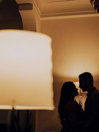 A romantic silhouette of the couple, creatively framed by a lampshade in the foreground. This intimate indoor shot uses ambient light to create a warm and moody atmosphere.