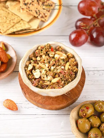 A bowl of savoury olive and nut dip, styled with crackers and grapes. The bright, airy lighting and white wooden background give it a fresh, Mediterranean vibe.