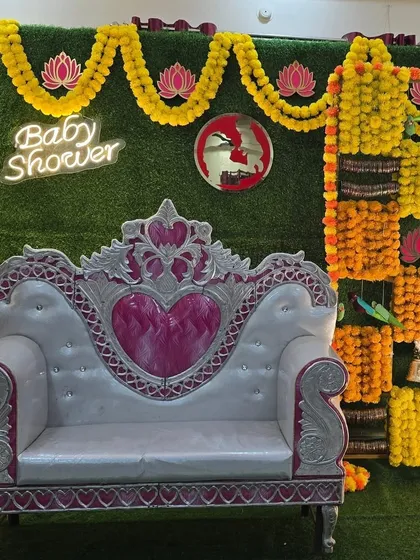 A beautiful and traditional baby shower setup combining classic and contemporary elements. The backdrop features a vibrant checkerboard pattern of marigold flowers on a green wall, complemented by a modern neon sign and an elegant silver and pink throne.