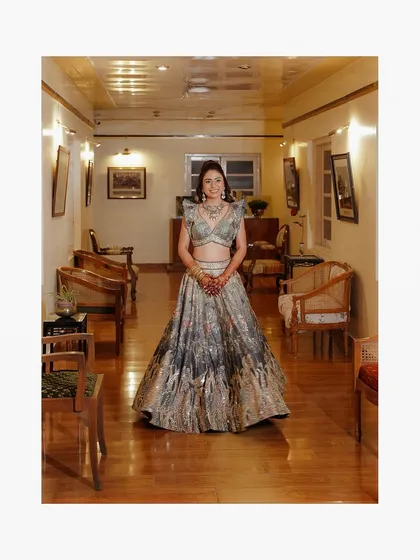 A full-length portrait of a woman in a stunning silver and grey lehenga, standing in a warmly lit, wood-paneled corridor.