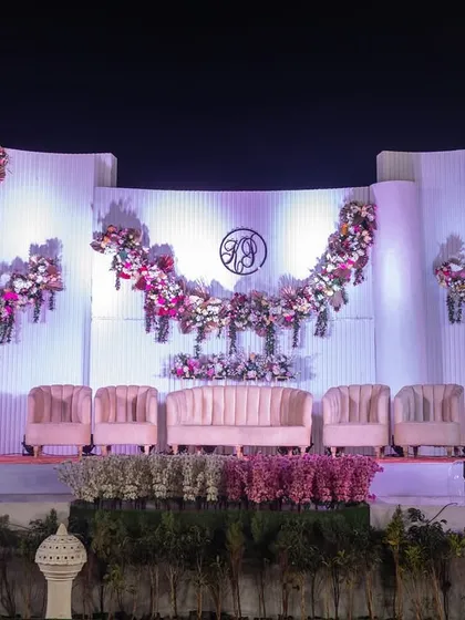 A grand reception stage with a unique, curved white backdrop and cascading floral arrangements. The soft pink seating and lighting create a romantic and elegant atmosphere.