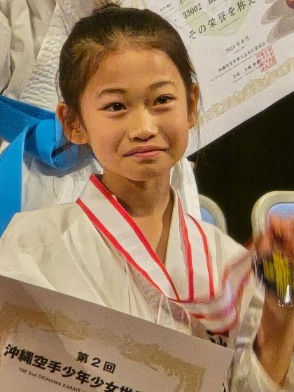 A young student's proud smile as she holds her certificate and medal from the Okinawa tournament. This is a moment of pure achievement and joy.