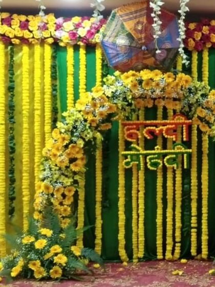 A combined "Haldi Mehandi" backdrop with a floral arch made of sunflowers and marigolds. The green and yellow drapes are complemented by Rajasthani umbrellas, creating a vibrant and traditional look.