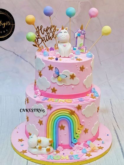 A magical two-tier unicorn-themed cake in a lovely shade of pink. It's decorated with fondant clouds, stars, a rainbow, and balloon cake pops for a whimsical first birthday.