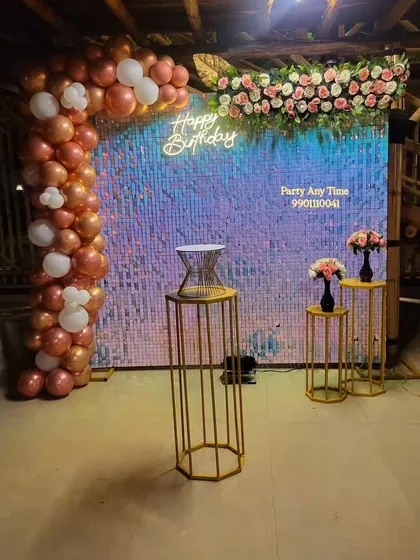 I can set up decorations at any venue, including resorts in Kanakapura. This surprise birthday features a beautiful shimmer wall, a rose gold and white balloon arch, and artificial flower arrangements for an elegant touch.