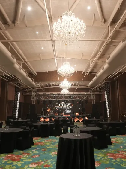 A wide view of the ballroom set up for a Sangeet night, with elegant black table settings, grand chandeliers, and a professional stage ready for performances.