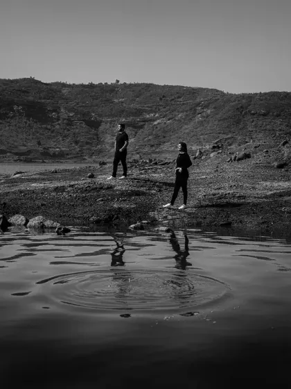 A wide, atmospheric black and white shot of the couple by a lake. The ripples in the water add a dynamic element to the otherwise still and serene landscape.
