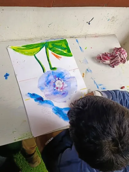 A close-up of the watercolor process. The student is carefully adding layers of blue to create depth in the flower petals.