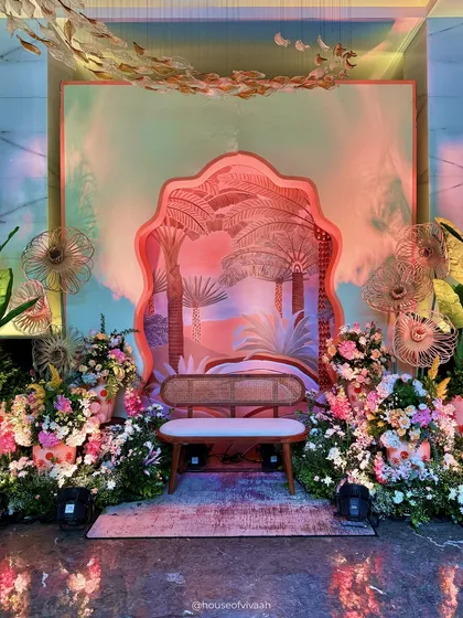 A whimsical photo backdrop for the Welcome Lunch, featuring a layered, scalloped arch with a palm tree motif, surrounded by lush florals and artistic fan-like structures.