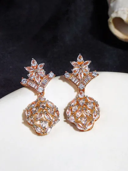 Intricate rose gold earrings styled on a white surface with a dark background. The lighting is carefully controlled to highlight the sparkle of the stones.