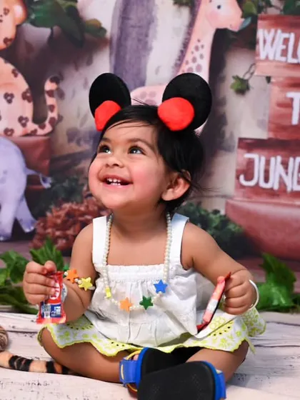 A fun shot from a Jungle Safari photoshoot, where the birthday girl is styled with Mickey Mouse ears. The backdrop includes a variety of animal characters like elephants, leopards, and monkeys.