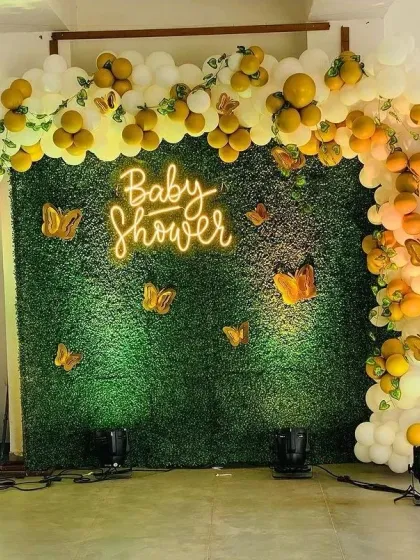 A chic and modern baby shower setup featuring a lush green foliage wall as the main backdrop. The design is highlighted by a custom 'Baby Shower' neon sign, a flowing balloon arch in white and yellow, and gold butterfly accents for an elegant touch.