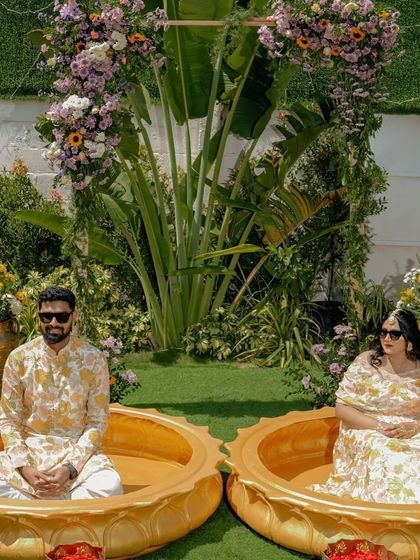 The bride and groom seated in their custom lotus-shaped tubs, ready for the Haldi ceremony to begin. The surrounding greenery and floral arch add to the fresh, garden-party feel of the celebration.