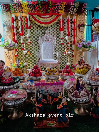 A grand Seemantham setup featuring a silver throne and a checkerboard backdrop in red, yellow, and white. The stage is filled with an elaborate display of traditional offerings and dolls.