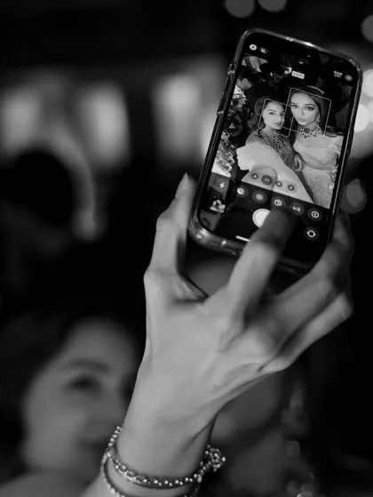 A guest takes a selfie with the bride in the background. This black and white photo captures a modern wedding moment, focusing on the layers of action and the joy of sharing the day.