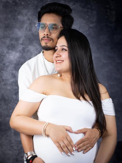 A classic studio portrait of an expecting couple. They stand together against a mottled grey backdrop, creating a timeless and sophisticated look.