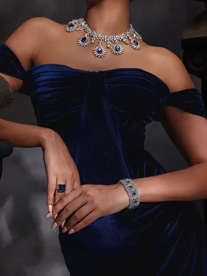 A shot of Purab Paschim's blue sapphire floral motif necklace, bracelet, and ring set against a rich blue velvet dress.