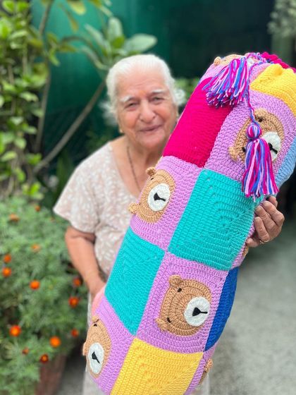 A colorful, bear-themed bolster pillow held by Dadi. The bright squares and cute bear faces make it a fun and functional piece of decor.