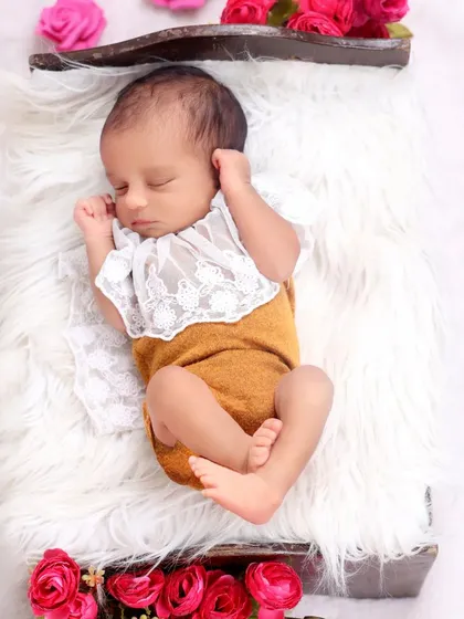 This full-body shot displays a beautiful outfit with a lace collar, set against a plush white fur rug. The addition of fresh pink roses adds a gentle pop of color to this classic newborn setup.